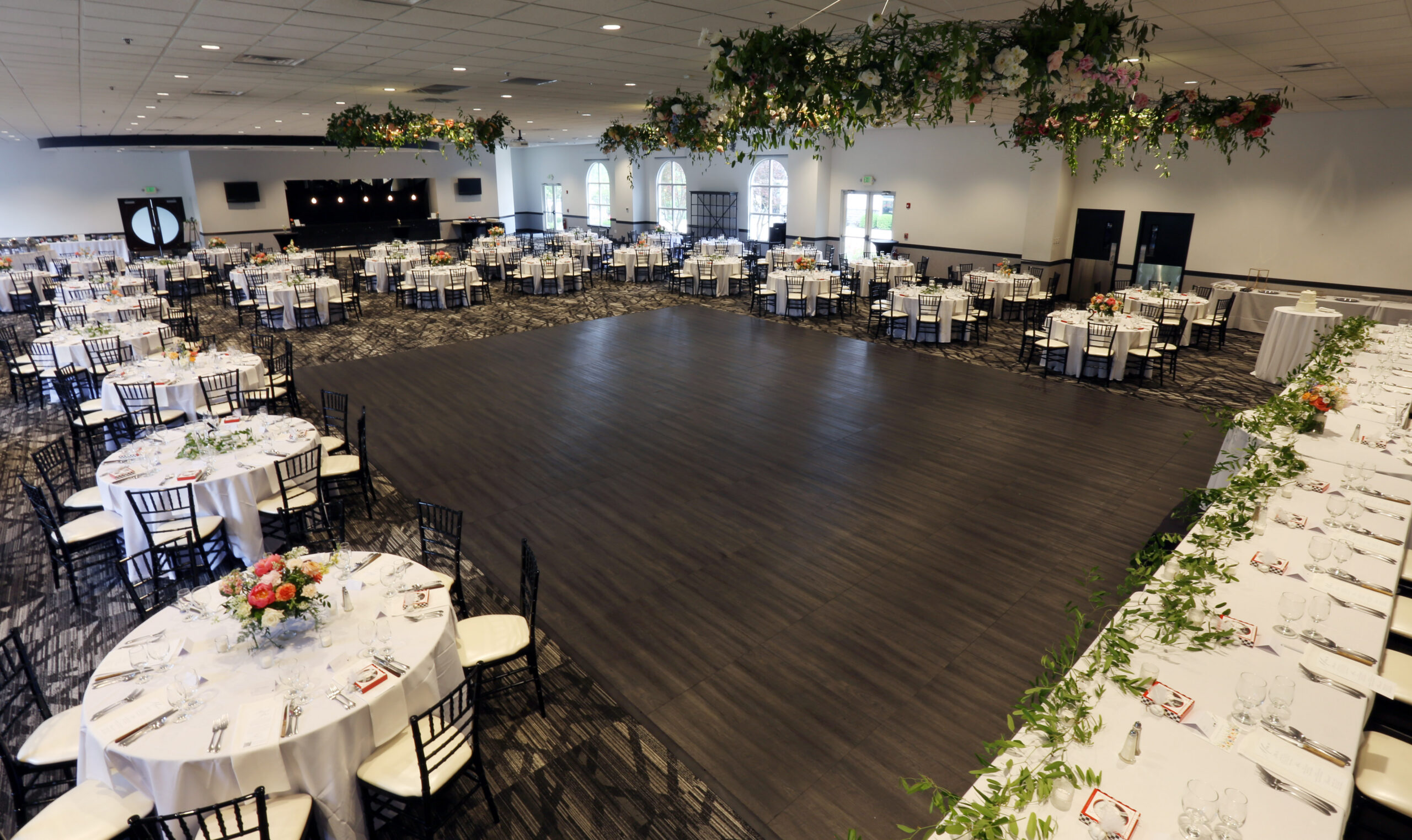 Elegant wedding reception ballroom at Ceruti’s in Fort Wayne, featuring round guest tables, floral decorations, a spacious dance floor, and greenery suspended above.